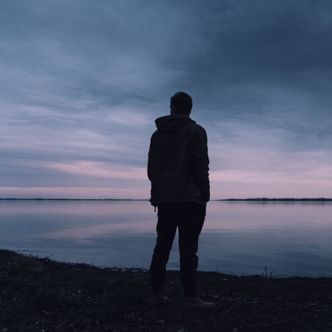 A man standing alone in front of a placid lake with a dark cloudy sky