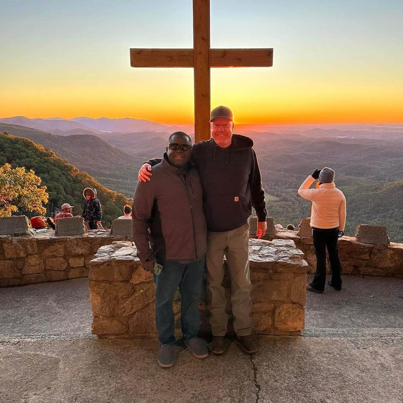 Men standing hugging each other before a cross with a sunset and mountain vista in the background
